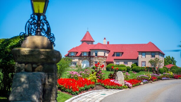 A large, red-roofed building with a stone facade, surrounded by well-maintained gardens and a cobblestone driveway. - The Mansion at Ocean Edge (Brewster)