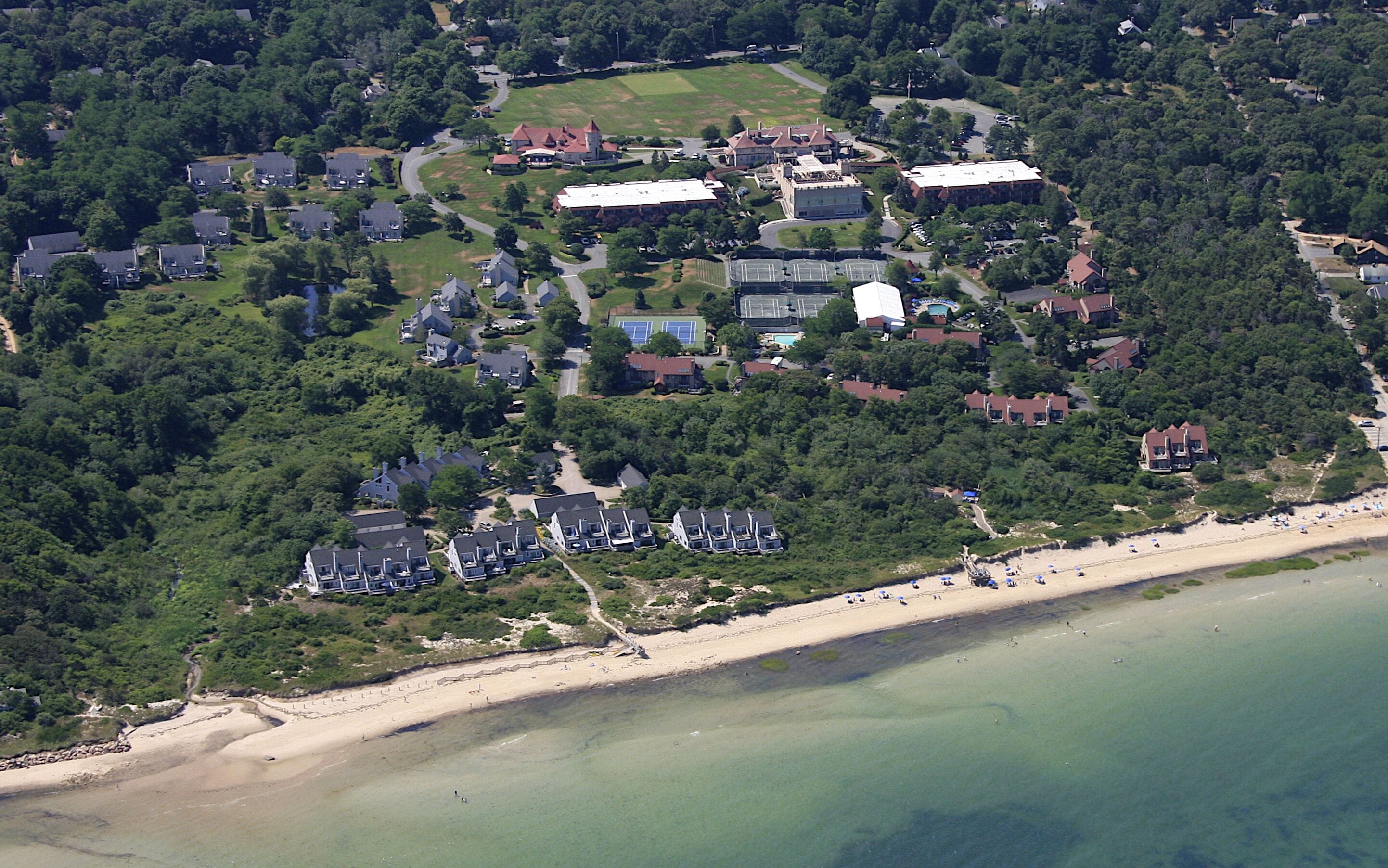 Vue aérienne d’un quartier résidentiel situé sur la côte, avec des maisons, une plage et un grand bâtiment doté d’un court de tennis.