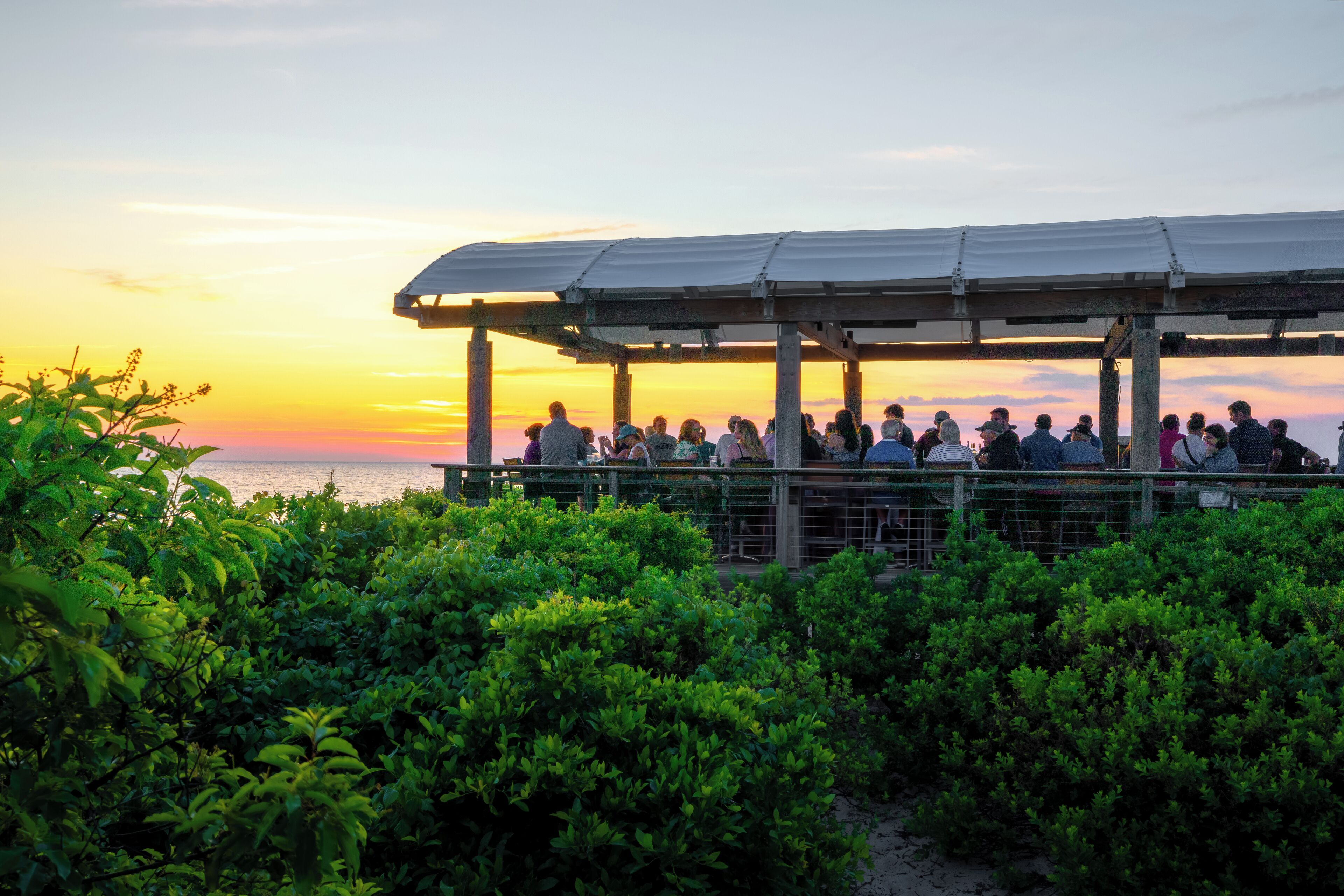 A covered outdoor seating area with a view of the sunset over the ocean.