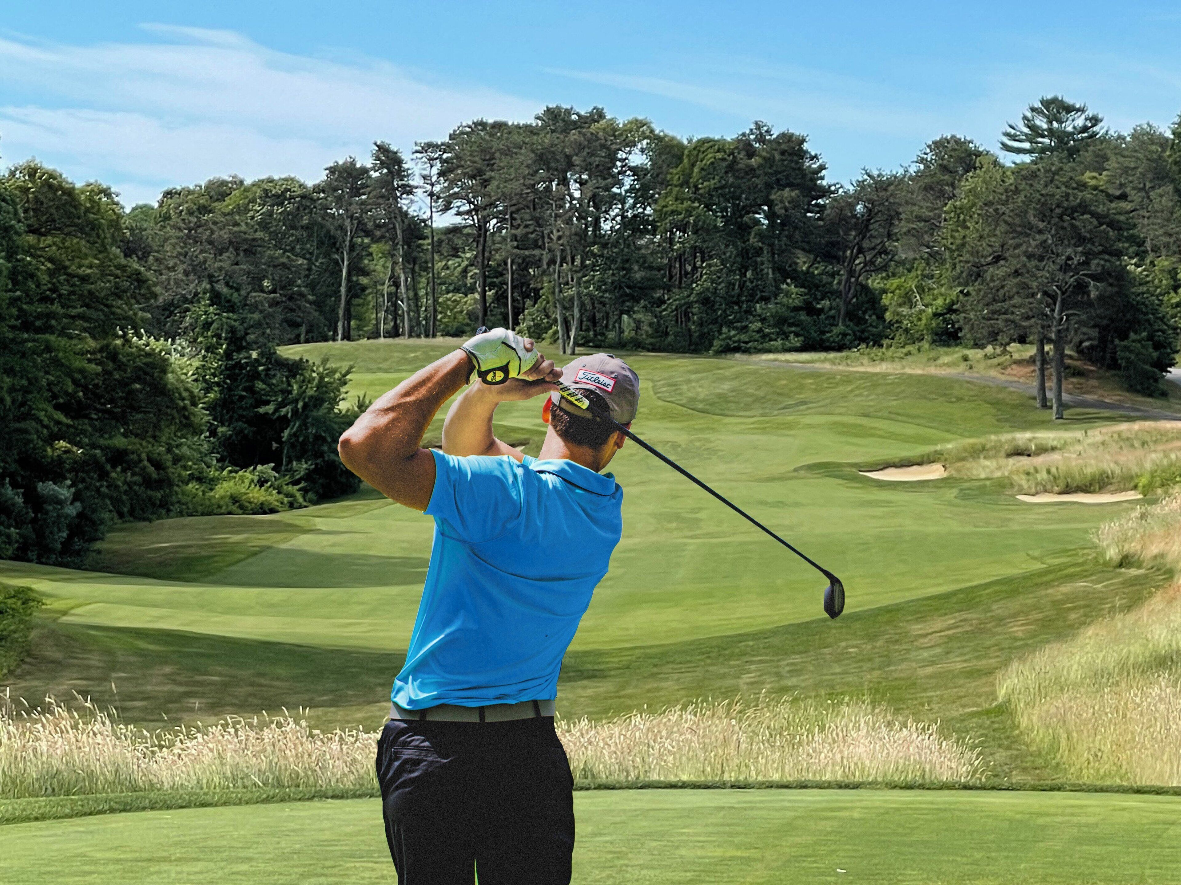 A golfer in a blue shirt and black pants preparing to swing on a golf course with a clear sky and trees in the background.