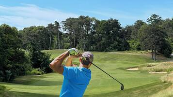 A golfer in a blue shirt and black pants preparing to swing on a golf course with a clear sky and trees in the background.