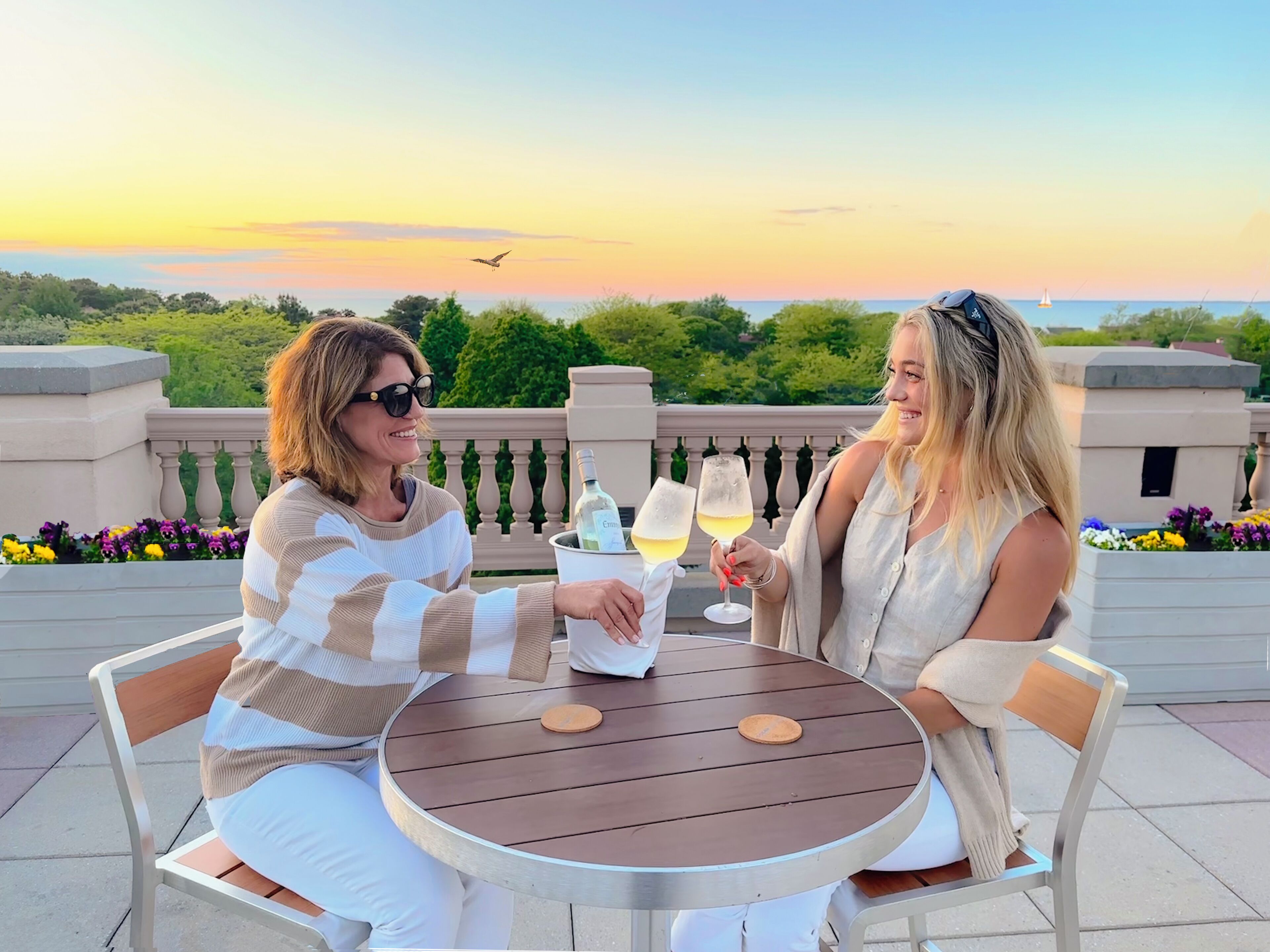 Duas pessoas sentadas em uma mesa redonda com uma garrafa de água e dois copos de vinho, ao ar livre, com vista para o mar e um pôr do sol ao fundo.
