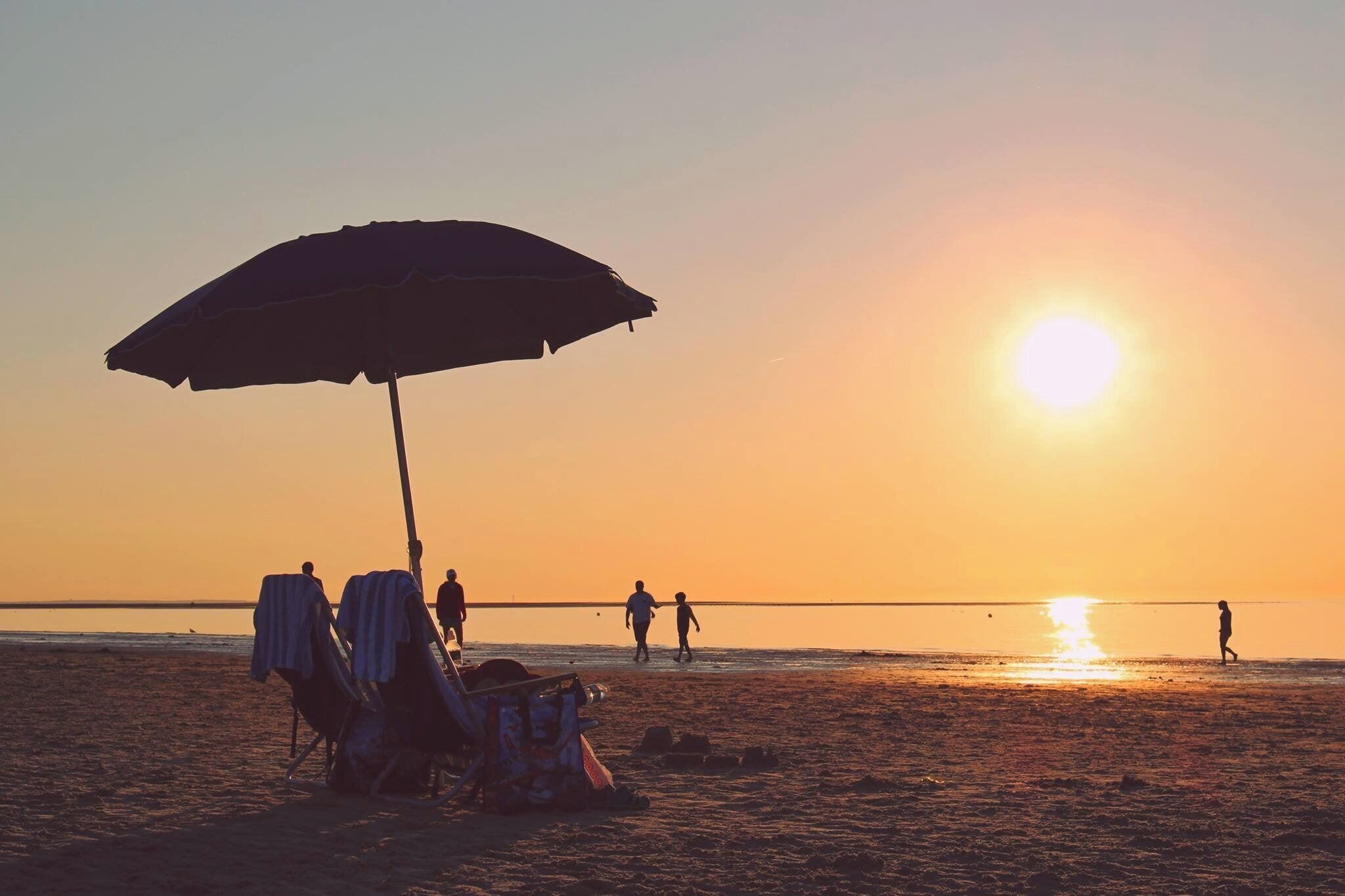 A beach scene with a large umbrella, beach chairs, and people walking along the shore.