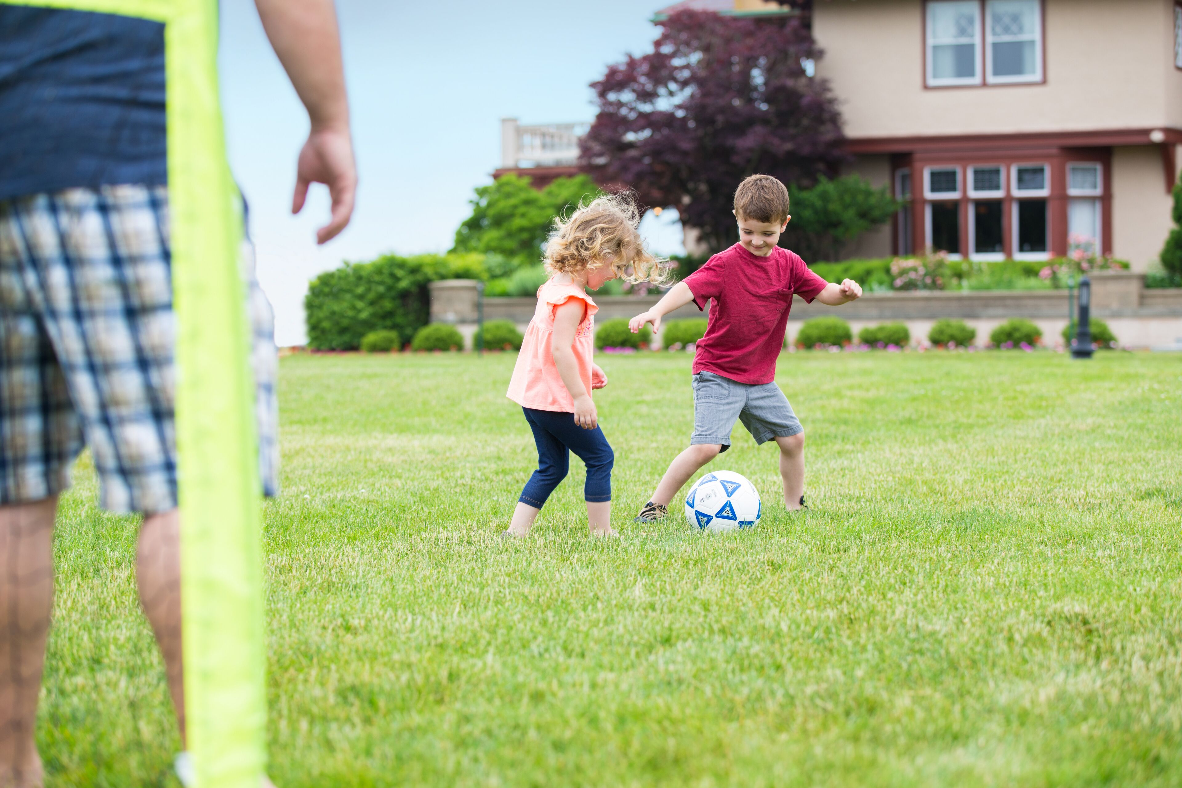 Deux enfants jouent au ballon sur un terrain herbeux, avec une maison en arrière-plan.