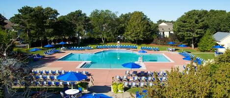 A pool area with multiple lounge chairs and umbrellas, surrounded by trees and a building in the background.