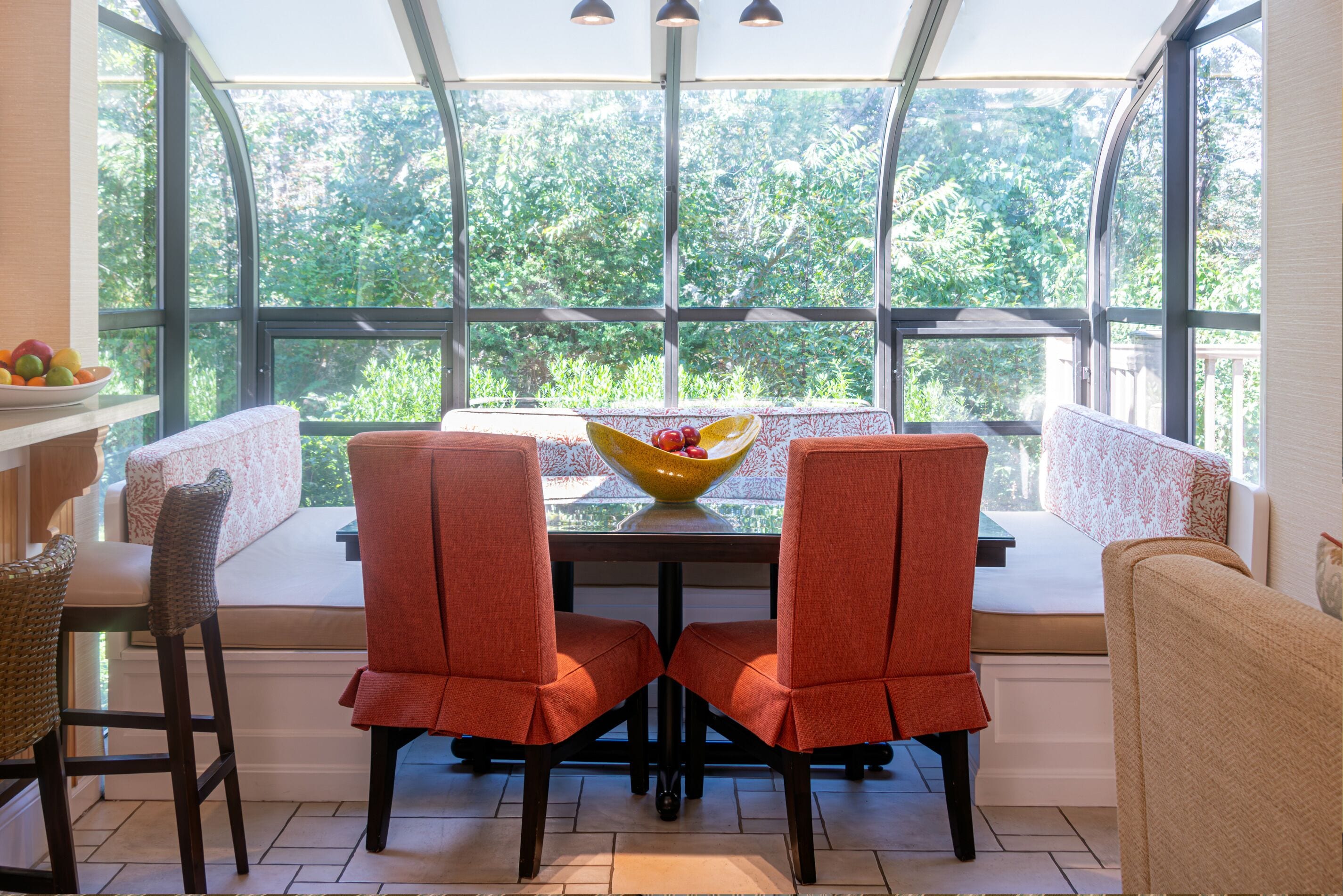 A dining area with a table and chairs, a bowl of fruit, and a view of greenery through large windows.