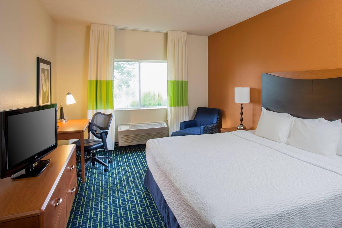 A hotel room with a bed, a desk with a computer, a chair, a TV, a lamp, and a window with curtains.