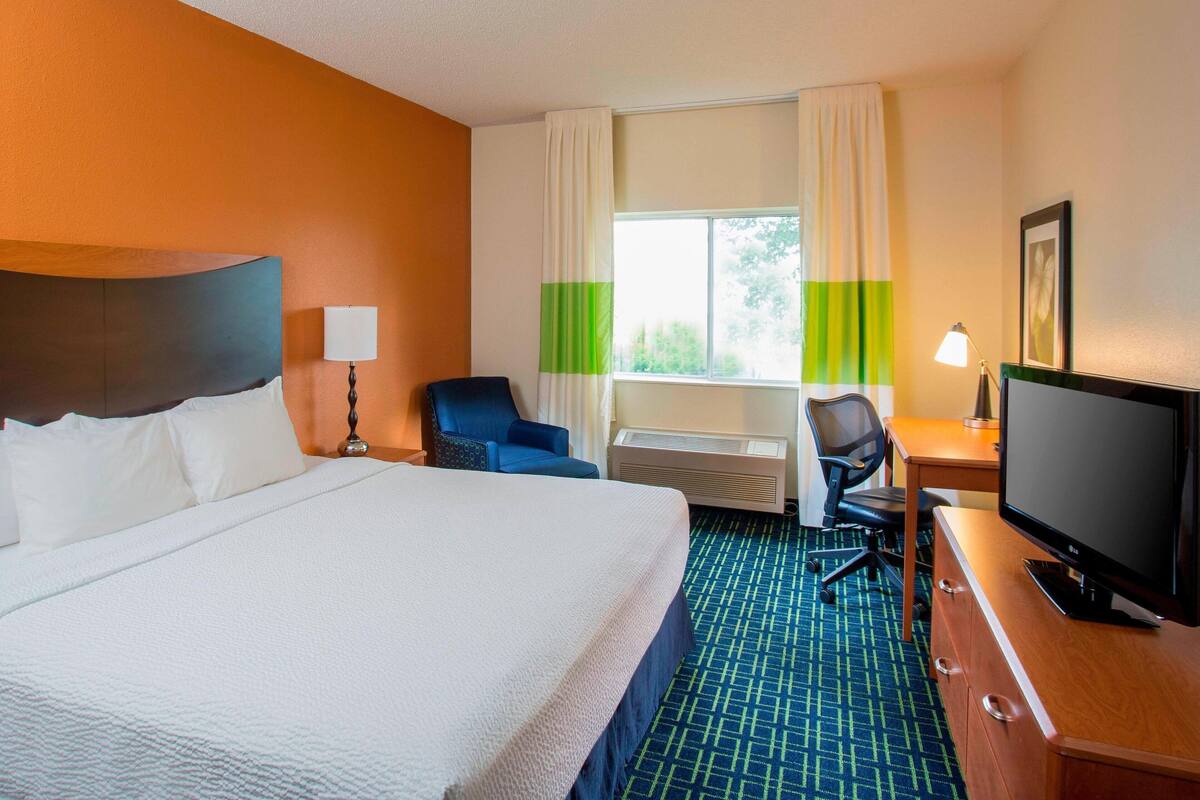 A hotel room with a large bed, a desk with a computer, a chair, a television, and a window with curtains.