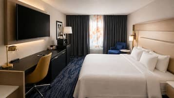 A hotel room with a bed, desk, chair, TV, and a view of the outdoors.