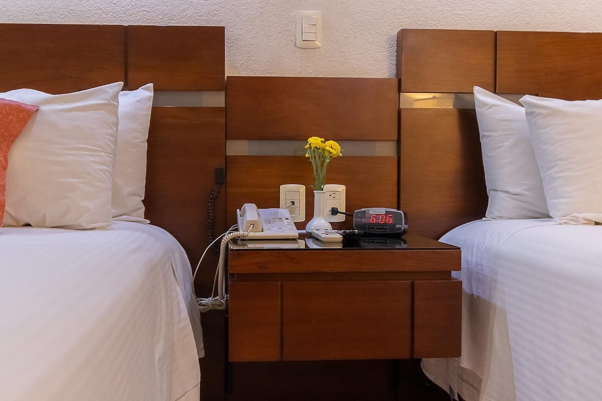 Une chambre d’hôtel avec deux lits, une table de chevet avec une horloge, un téléphone et un vase avec des fleurs.