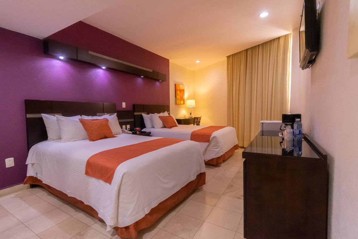 A hotel room with two beds, a desk, a TV, and a window with curtains.
