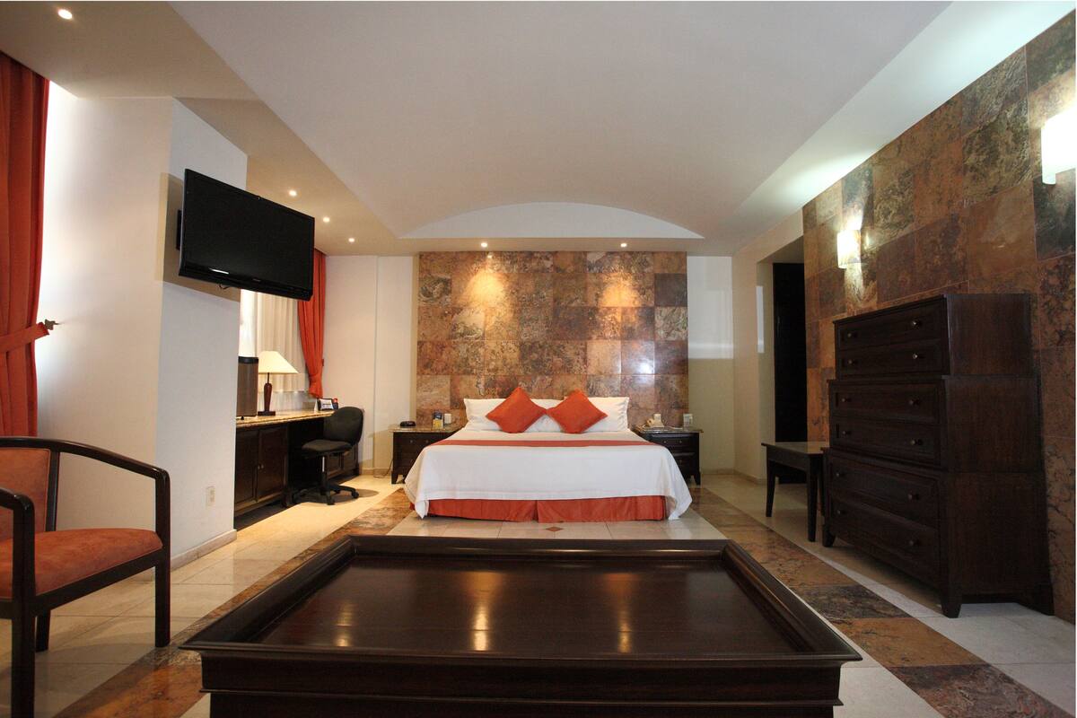 A hotel room with a large bed, a flat-screen TV mounted on the wall, a desk with a lamp, and a dresser.