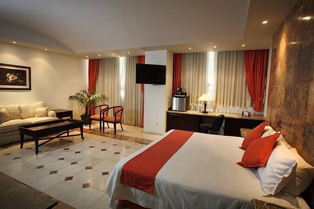 A hotel room with a bed, a desk, a chair, a sofa, a TV, and a plant.