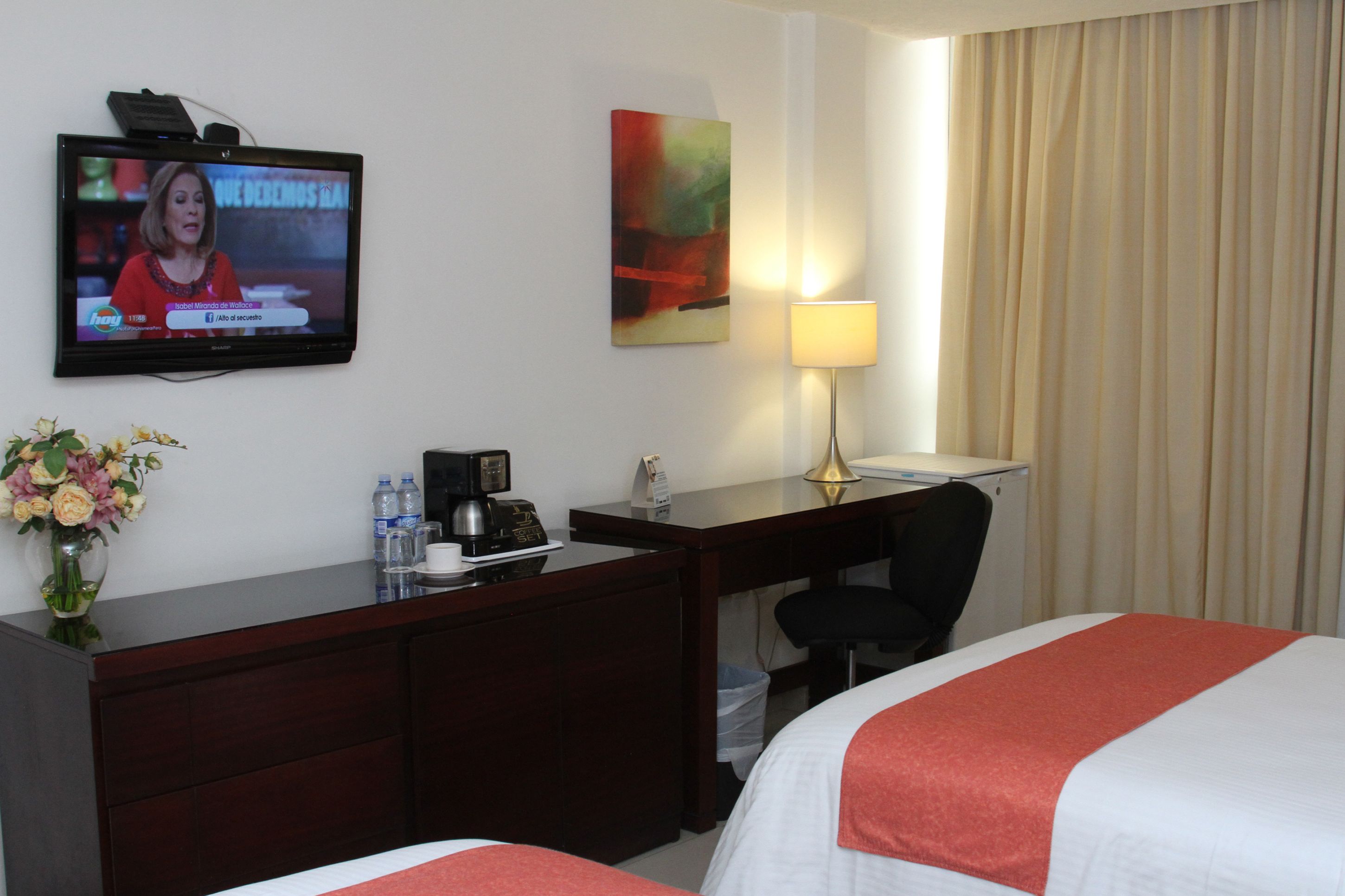 Habitación de hotel con una televisión en la pared, un escritorio con lámpara, una silla y dos camas.