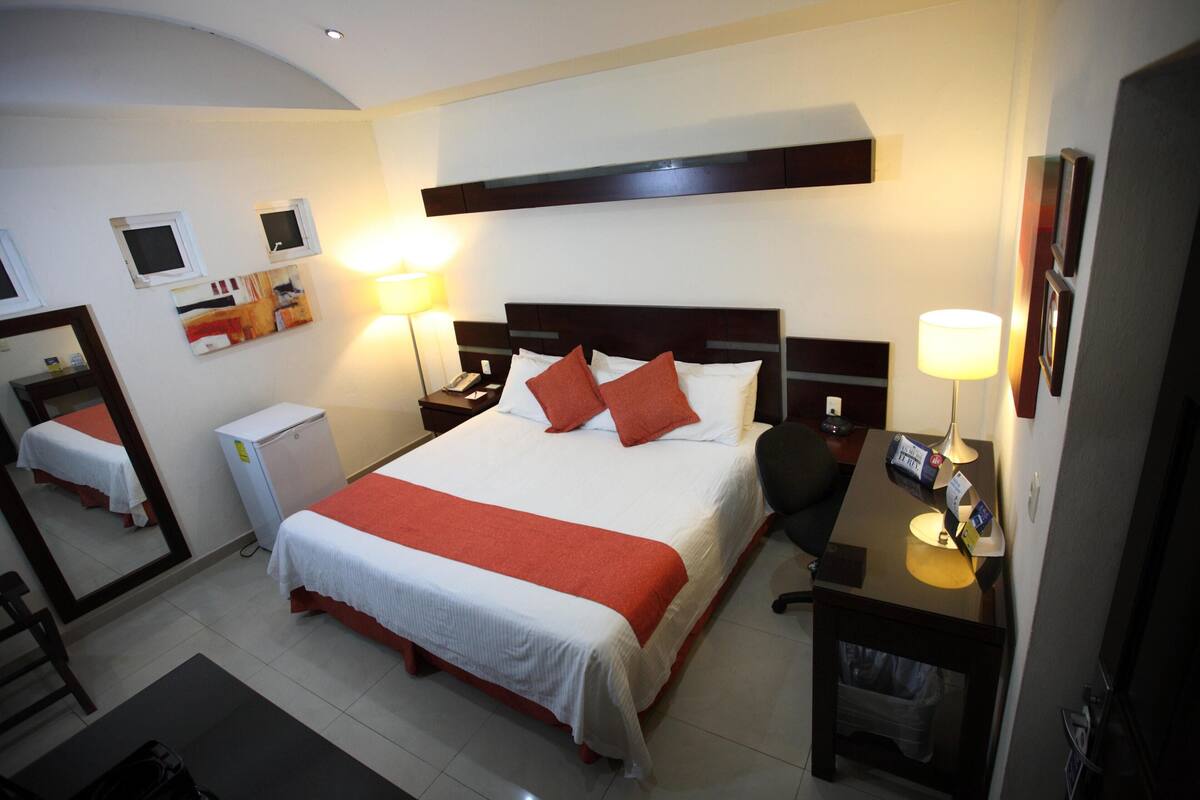 A hotel room with a large bed, bedside tables, a desk, a chair, a mirror, and a small refrigerator.