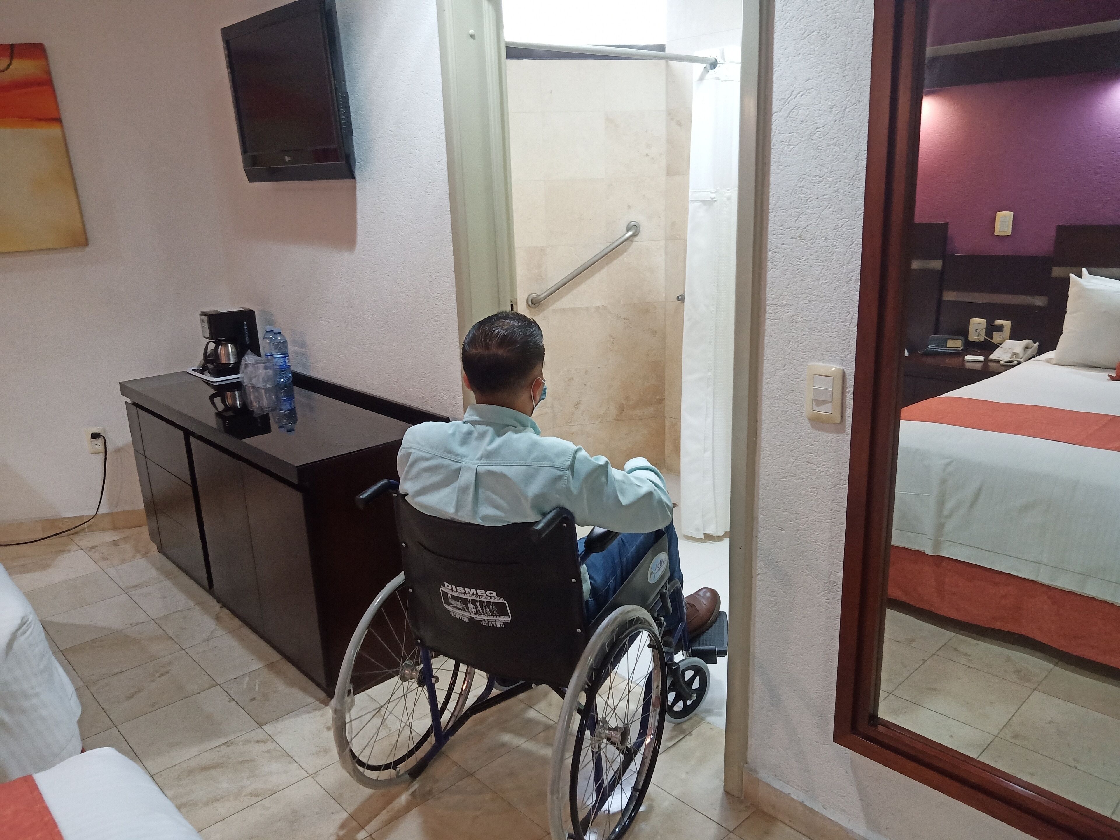 A wheelchair-accessible room with a bed, a desk with a coffee maker and water bottles, a TV on the wall, and a bathroom visible through a glass door.