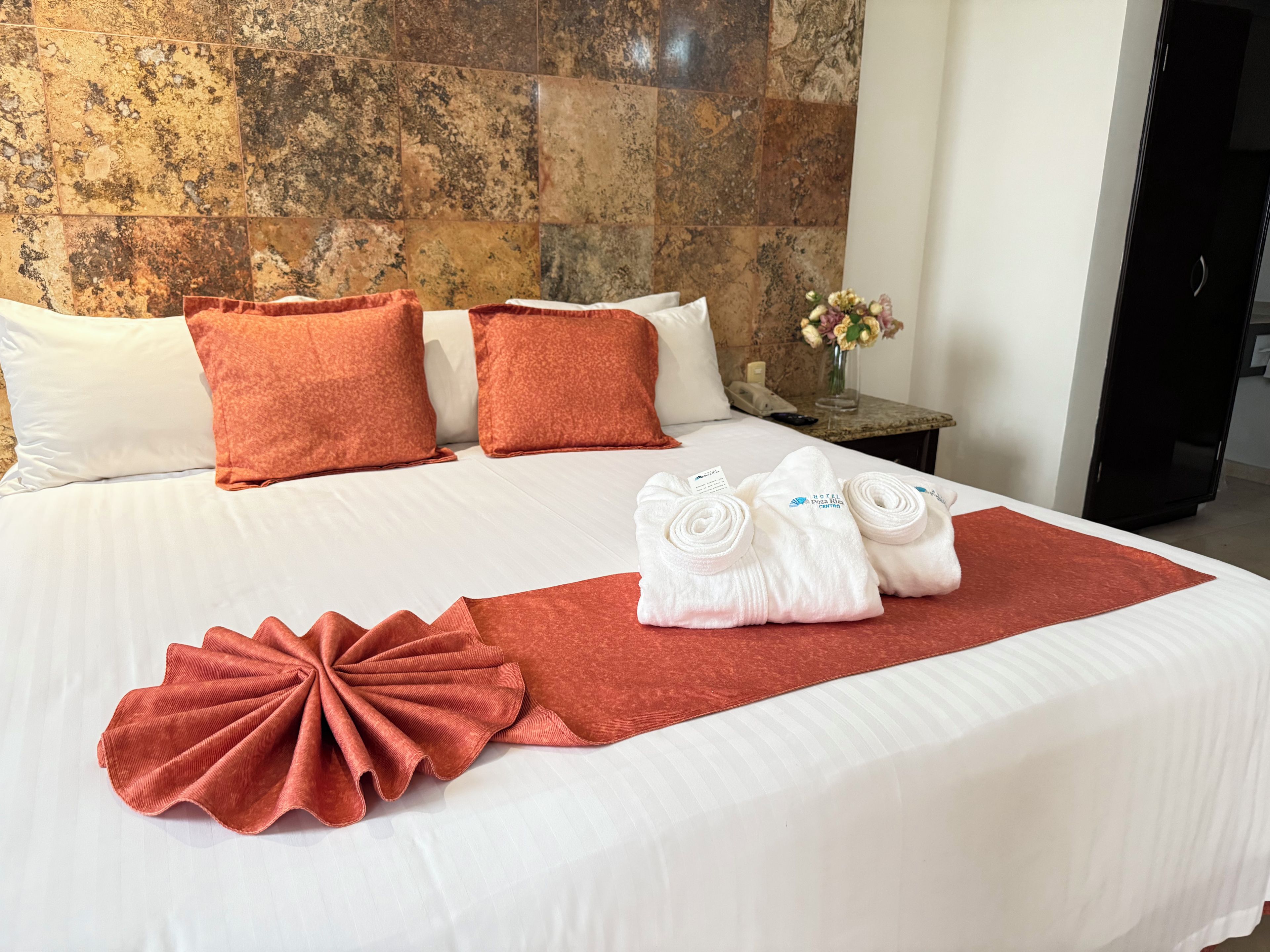 Habitació d'hotel amb un llit gran, almoinos blancs i taronja, una manta vermella i tovalloles ben esteses sobre el llit.