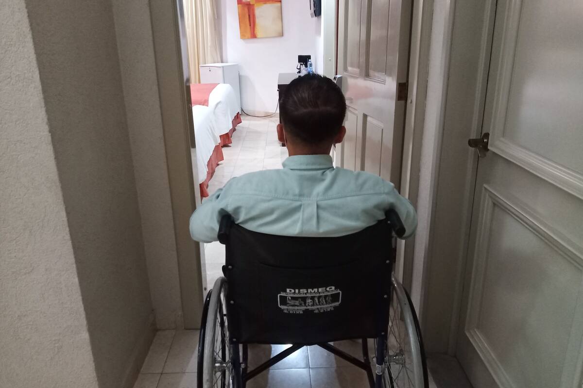 A person in a wheelchair in a narrow hallway with a door on the right and a bed with a red blanket on the left.