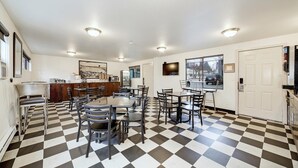 A dining area with checkered flooring, multiple tables and chairs, a kitchen area with a refrigerator, and a large window offering a view of the outside. - The Grove West Seattle Inn (Seattle)