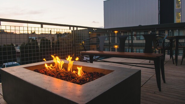 A rooftop terrace with a square fire pit, wooden deck, metal chairs, and a view of a modern building. - The Grove West Seattle Inn (Seattle)