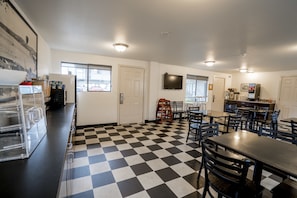 A restaurant with a checkered floor, multiple tables and chairs, a kitchen area, and a television mounted on the wall.