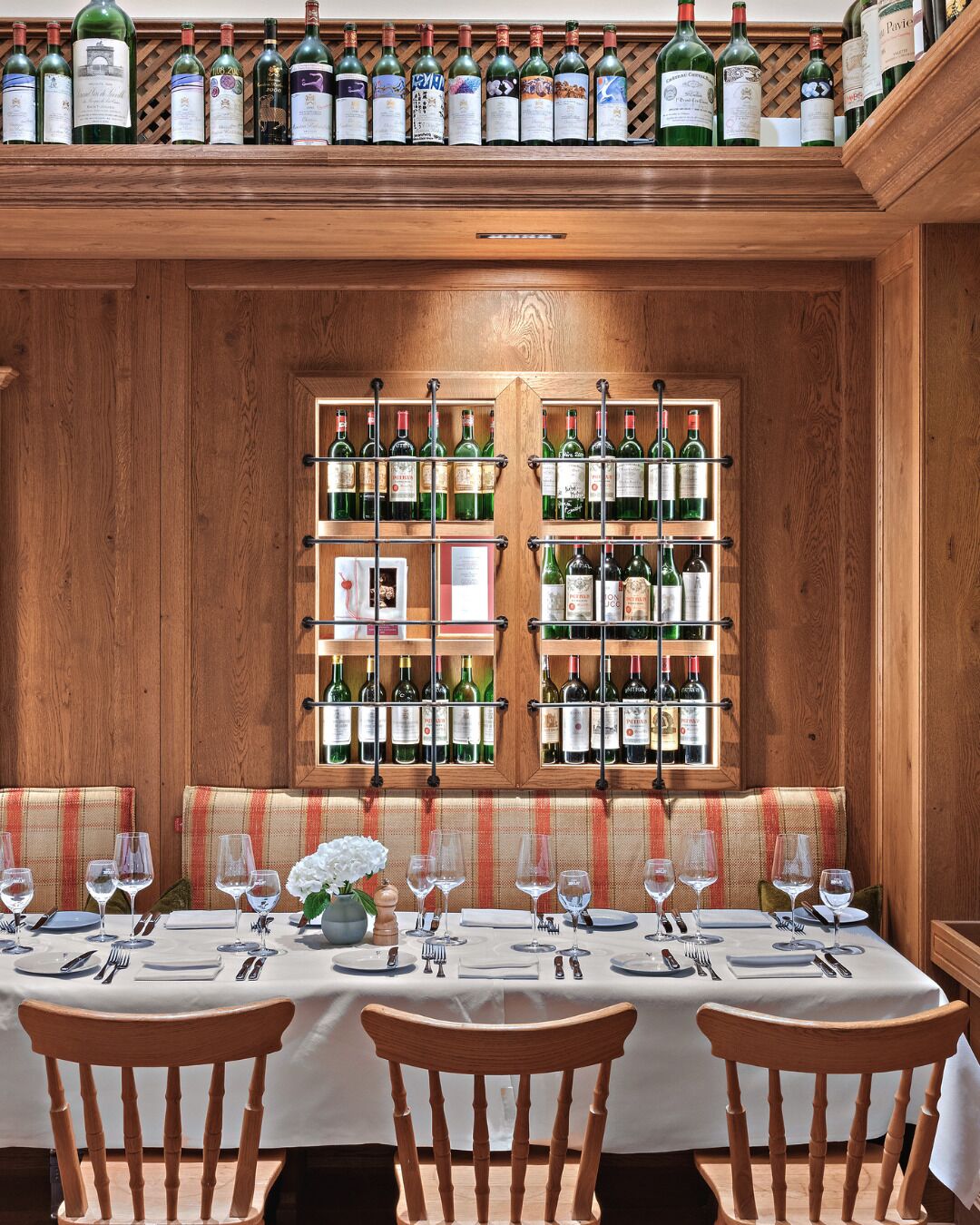 L’intérieur d’un restaurant présente une longue table en bois dressée pour le repas, de nombreuses bouteilles de vin exposées sur des étagères et un banc à carreaux.