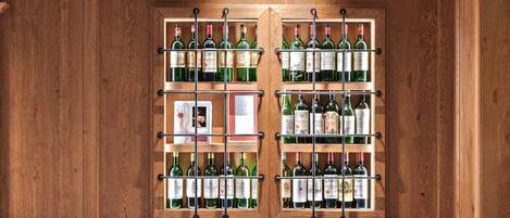 L’intérieur d’un restaurant présente une longue table en bois dressée pour le repas, de nombreuses bouteilles de vin exposées sur des étagères et un banc à carreaux.