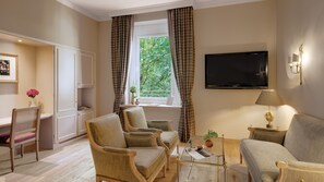 A living room with a sofa, two armchairs, a coffee table, a TV mounted on the wall, a desk with a chair, and a window with curtains.