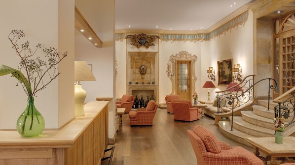 A grand foyer with a fireplace, checkered armchairs, a staircase, and a green vase with branches. - Hotel Excelsior München (Munich)
