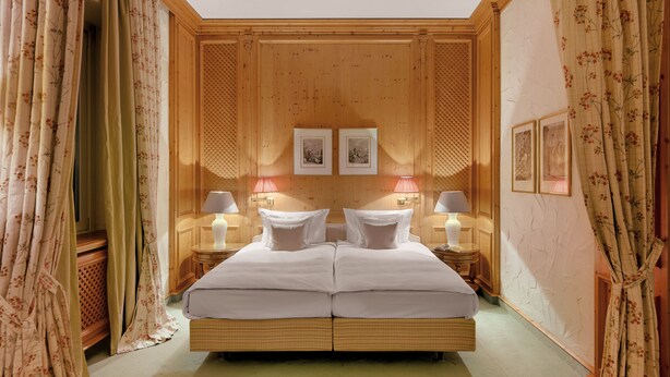 A double bed with two nightstands, each with a lamp, in a room with wooden paneling and floral curtains. - Hotel Excelsior München (Munich)