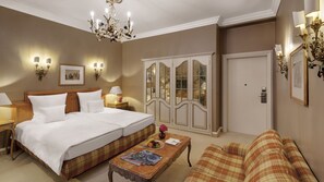 A hotel room with a large bed, a sofa, a coffee table, a chandelier, and a wardrobe with glass doors. - Hotel Excelsior München (Munich)