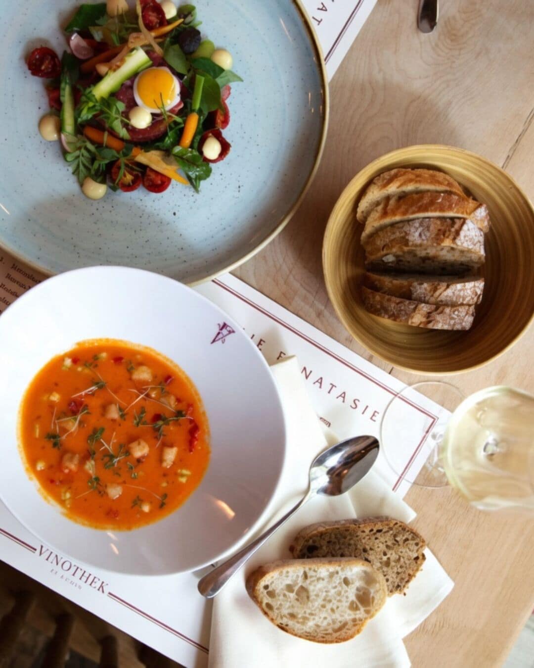 A table with a bowl of soup, a plate of salad, a basket of bread, and a glass of white wine.