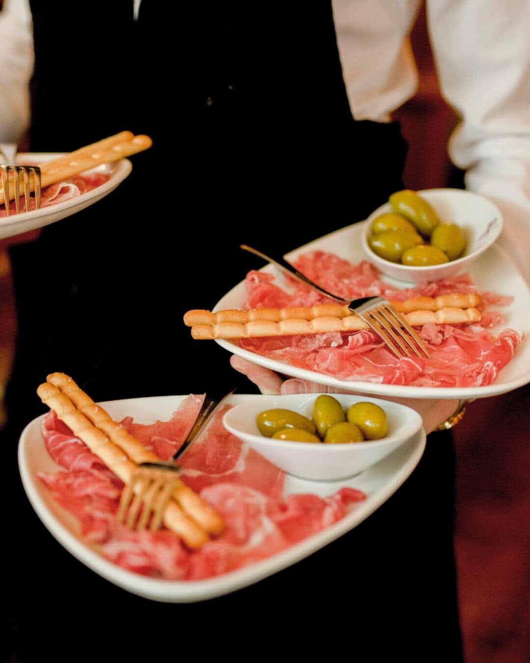 A person holding plates with sliced meat, olives, and crackers.