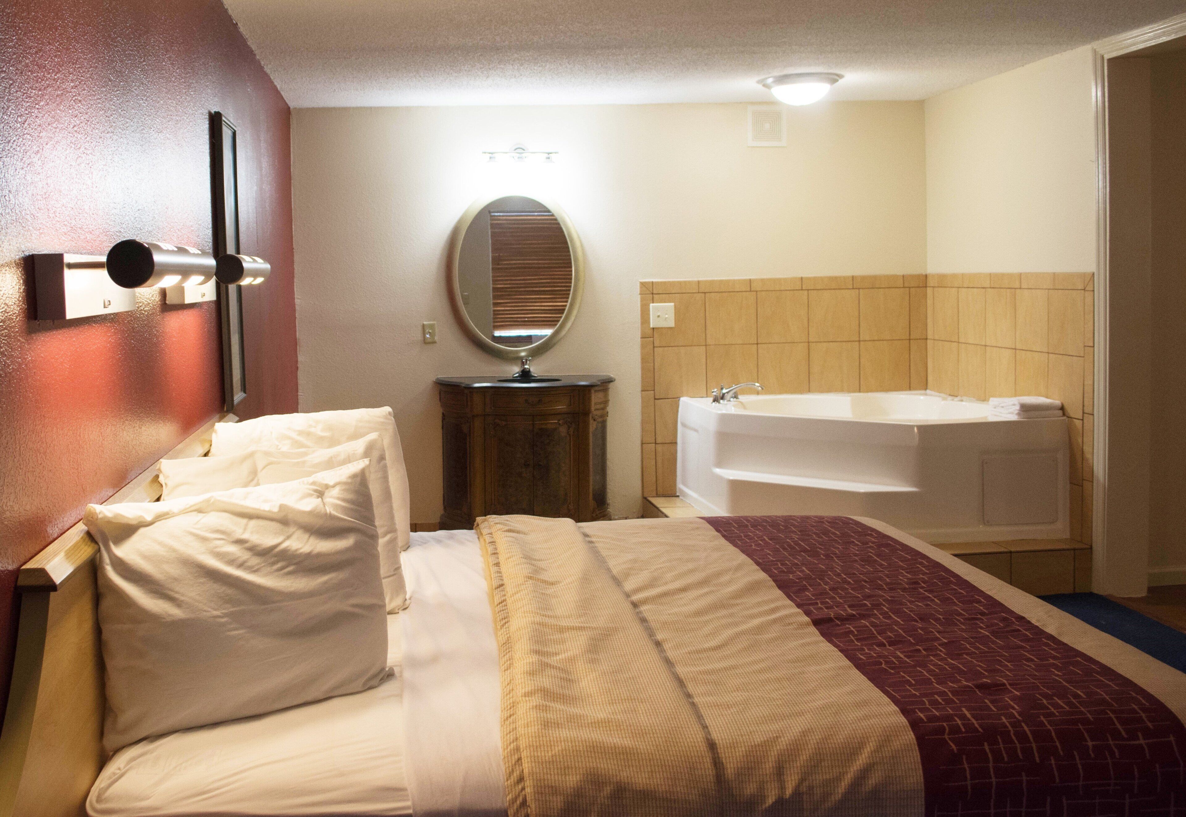 A hotel room with a bed, a bedside table, a mirror, a wall-mounted light, and a bathtub.