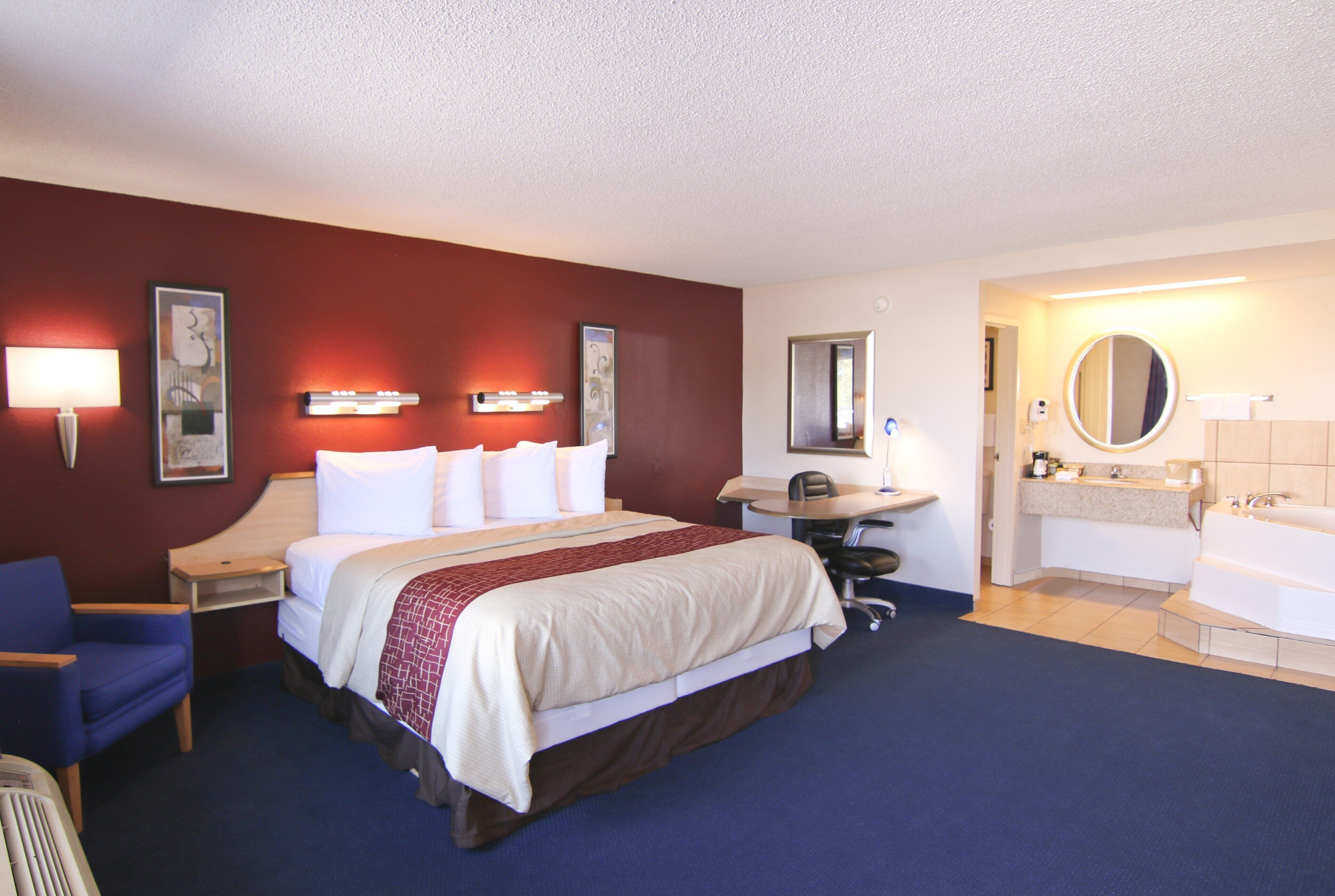 A hotel room with a large bed, a desk, a chair, a desk lamp, a round mirror, a sink, and a bathtub.