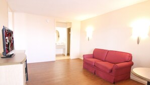 A room with a wooden floor, a red sofa, a small round table, and a TV on a stand. - Red Roof Inn Somerset, KY (Somerset)