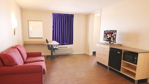 Une pièce de taille modeste, comprenant un canapé rouge, un bureau avec une chaise, un téléviseur et un four à micro-ondes.