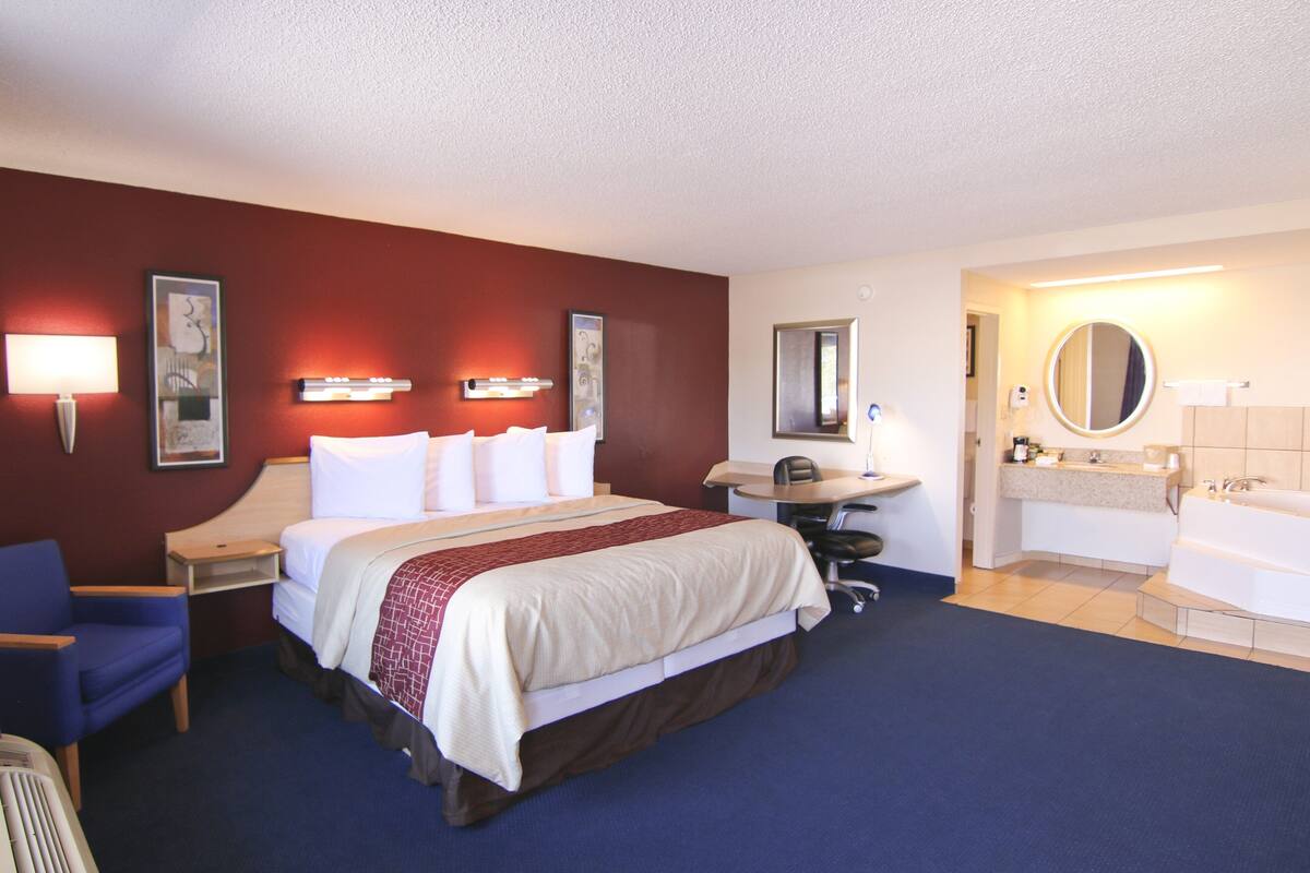 A hotel room with a large bed, a desk, a chair, a round mirror, a sink, and a bathtub.