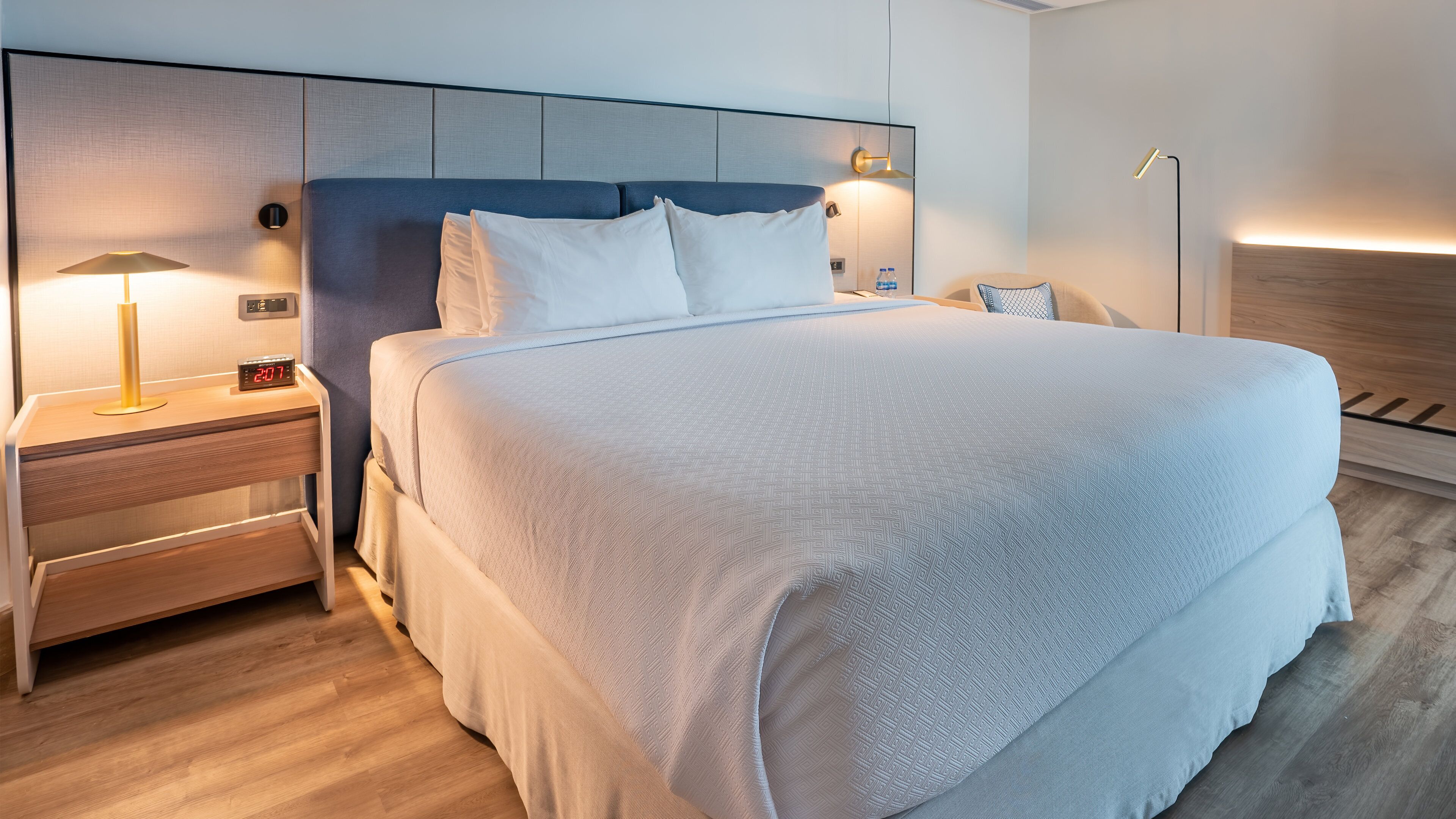 Une chambre d’hôtel moderne avec un grand lit, une table de chevet avec une lampe et une horloge numérique.