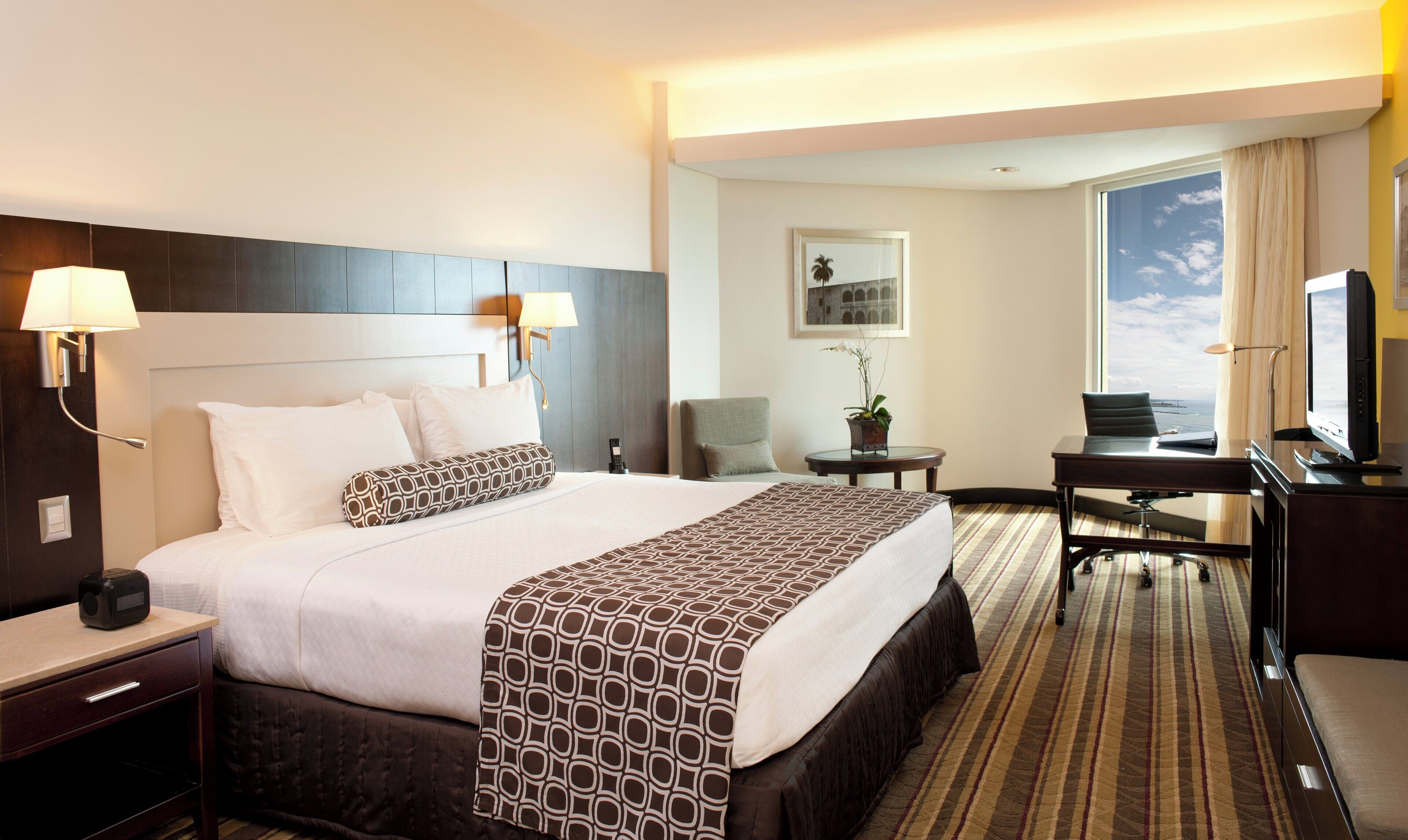 A hotel room with a large bed, bedside lamps, a desk with a television, and a view of the sky.