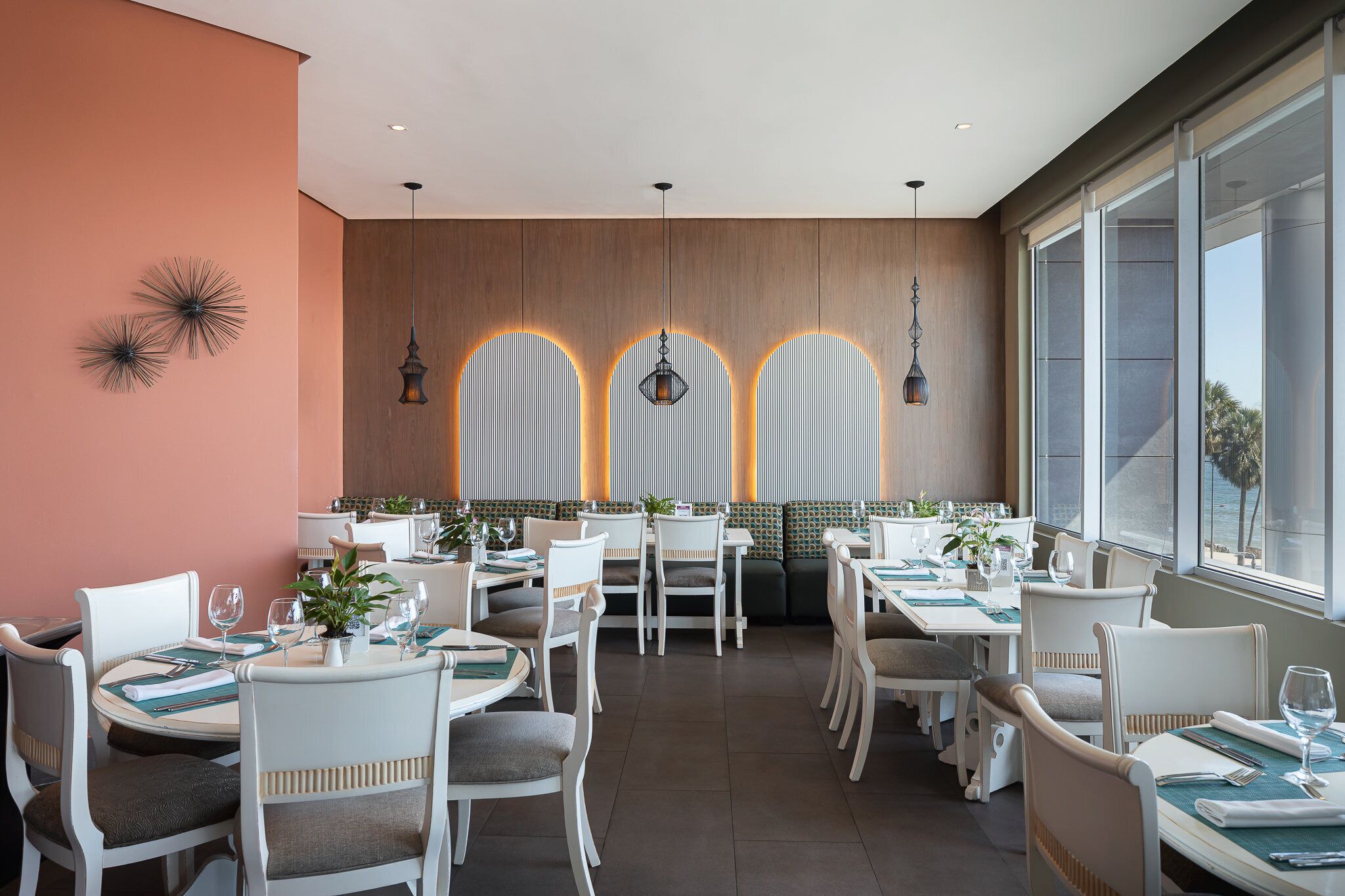 A restaurant with white chairs, tables set with plates, glasses, and cutlery. There are hanging lights and a wall with a geometric design.