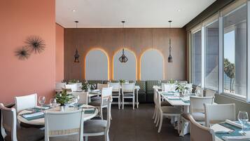 Un restaurant avec des chaises blanches, des tables dressées avec des assiettes, des verres et des couverts. On y trouve des lampes suspendues et un mur orné d’un motif géométrique.