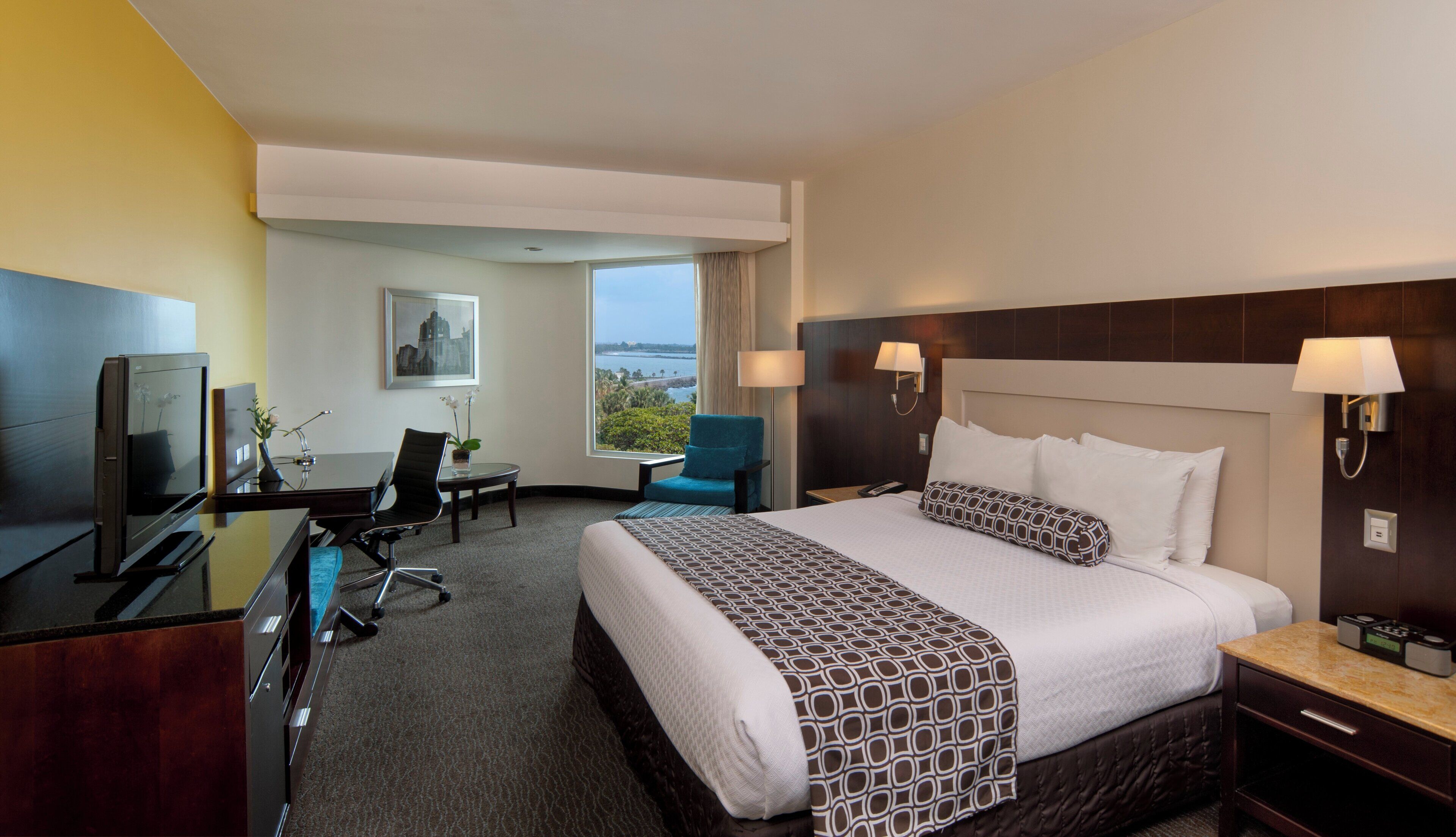 A hotel room with a large bed, a desk with a computer, a chair, a TV, and a window with a view of the ocean.