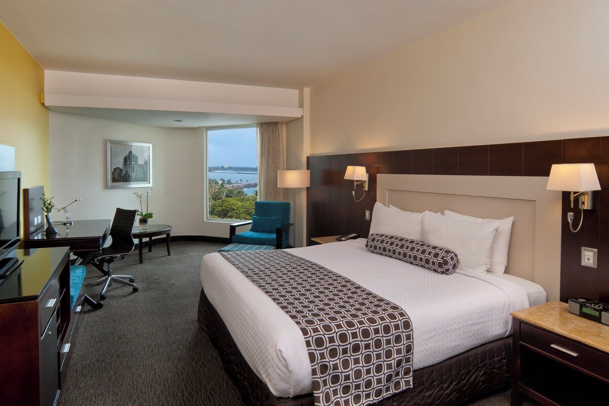 Ein Hotelzimmer mit einem großen Bett, einem Schreibtisch mit Computer, einem Sessel, einem Fernseher und einem Fenster mit Meerblick.