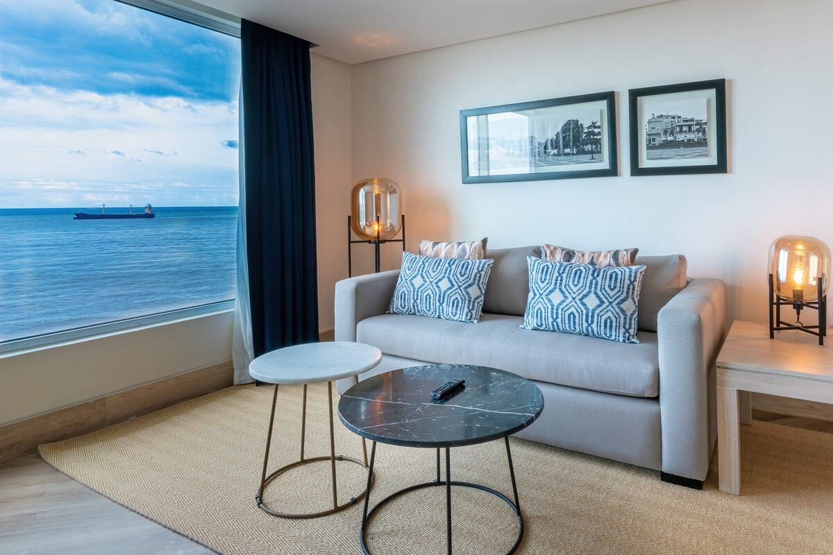 Ein modernes Wohnzimmer mit einer Couch, zwei gemusterten Kissen, einem kleinen runden Tisch und zwei Wandleuchten. Ein großes Fenster bietet Blick auf das Meer und ein Schiff.
