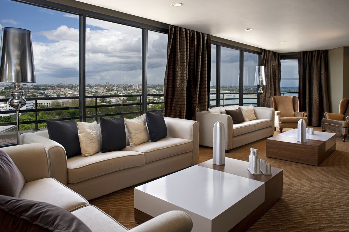 Ein geräumiges Wohnzimmer mit großem Fenster und Blick auf die Stadt, einer Sofagarnitur und zwei Couchtischen.