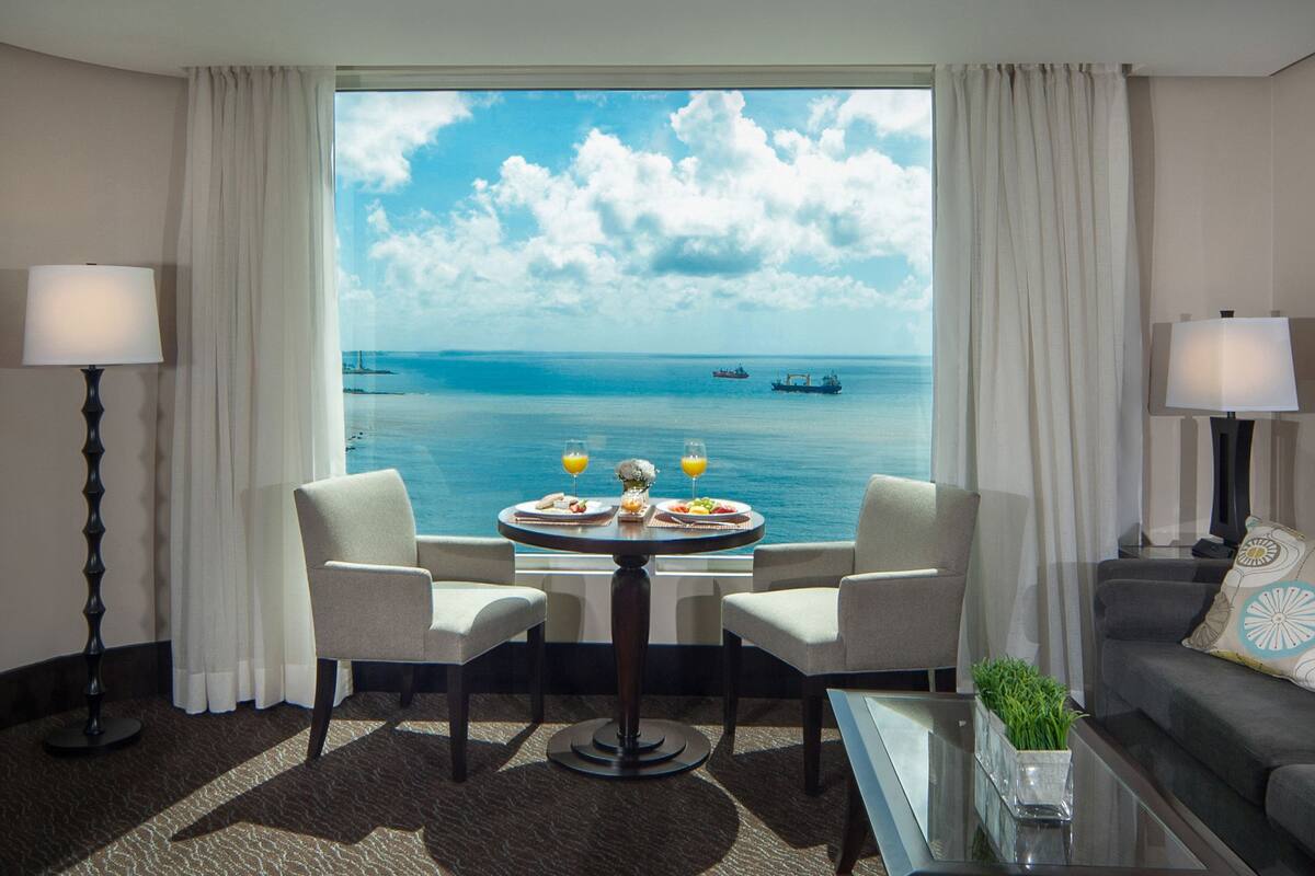 Une chambre d’hôtel dotée d’une grande fenêtre donnant sur la mer et les bateaux. On y trouve une table à manger avec des chaises, ainsi qu’une petite table basse agrémentée d’une plante.