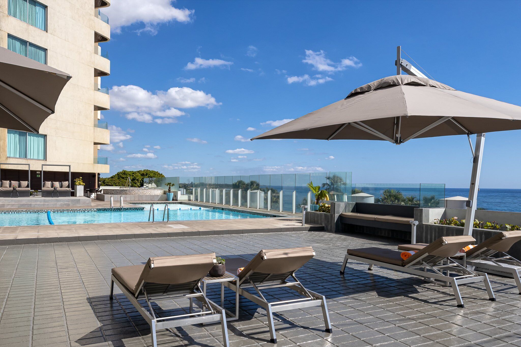 Un espace piscine aménagé sur le toit, avec des chaises longues et un grand parasol, offrant une vue imprenable sur un ciel bleu azur et l’océan.