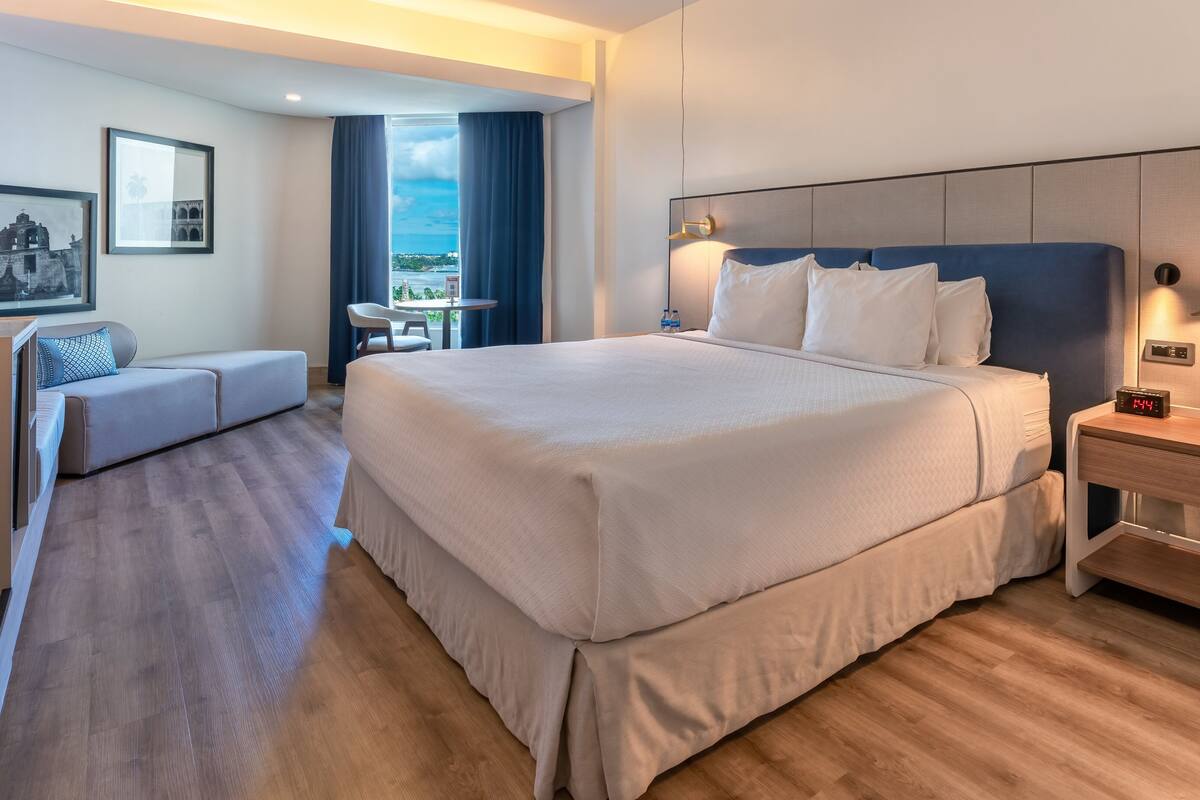 Une chambre d’hôtel avec un grand lit, une table de chevet avec une lampe, une chaise, une petite table avec une autre chaise, et une vue sur la mer.