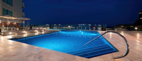 A swimming pool with a tiled deck and a metal handrail, surrounded by a building with large windows and outdoor seating.